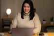 © Studio Marmellata - Positive young Arab woman in casual sweater sitting at table with laptop and working online on project from home during evening time