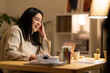 © Studio Marmellata - Positive young Arab woman in casual sweater sitting at table with laptop and working online on project from home during evening time