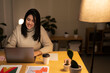 © Studio Marmellata - Positive young Arab woman in casual sweater sitting at table with laptop and making a video call on project during evening time