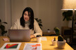 © Studio Marmellata - Positive young Arab woman in casual sweater sitting at table with laptop and making a video call on project during evening time