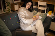 © Studio Marmellata - Concentrated Arab female in casual beige outfit and eyeglasses sitting on sofa with documents using laptop while working online at home