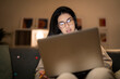 © Studio Marmellata - Arab female freelancer in casual wear and glasses typing information on laptop while working remotely sitting on sofa at home in evening