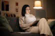 © Studio Marmellata - Arab female freelancer in casual wear and glasses typing information on laptop while working remotely sitting on sofa at home in evening