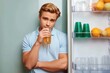 © Vladislav - An attractive young man in a blue polo shirt drinks juice while standing next to an open refrigerator filled with fruit and glasses. Healthy lifestyle.
