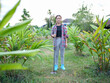 © buraratn - Young girl stands confidently lush garden, holding gardening tool. She wears plaid shirt over black top, paired with gray pants and blue boots. vibrant greenery surrounds her, creating serene