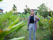 © buraratn - Young woman stands confidently lush green field, holding rolled up papers. She wears casual outfit with plaid shirt and gray pants, surrounded by vibrant plants and trees. scene conveys sense