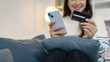 © PHOTO4U - A young woman is sitting on a sofa holding a smartphone and credit card, smiling as she makes an online transaction. She looks relaxed and confident while shopping at home.