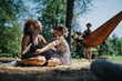 © qunica.com - Two friends share a joyful moment on a checkered blanket as others relax nearby; a hammock and guitar in the background create a casual, outdoor vibe during a sunny park gathering.