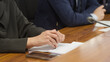 © slexp880 - Close-up of a lawyer or official in a suit holding a pen over documents at a wooden desk during a meeting; smartphone and mic visible, focus on hands and paperwork. Photo