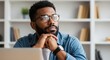 © Vasiliy - Thoughtful African American man with glasses in deep thought at his desk. Pensive young professional or student planning a project in a modern home office