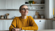 © SnapVault - Woman writing in a notebook, wearing glasses and a yellow sweater, smiling while looking away with a thoughtful and enthusiastic expression, planning her day and future success in a kitchen setting