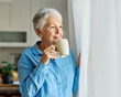 © Lumos sp - Portrait of a happy senior elderly woman holding a cup of coffee standing by window and relaxing at home
