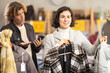 © JackF - Interested female customer choose new jacket or coat for winter season, man bored while waiting for woman and looking at smartphone in modern clothing shop