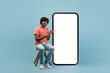 © Prostock-studio - A positive young Indian man with curly hair sits casually on a chair. He points at a large cell phone with a blank white screen, smiling happily against a blue studio background.