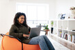 © Prostock-studio - A young woman with curly hair sits comfortably on an orange bean bag chair, smiling as she types on her laptop. The bright living room has modern decor and plants.