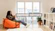 © Prostock-studio - A woman relaxes in a stylish living room, sipping from a mug. She sits in an orange chair, surrounded by light and modern decor, creating a cozy atmosphere perfect for unwinding.