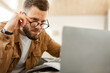 © Prostock-studio - A man with a beard sits at a desk in a modern workspace, leaning forward to closely examine the content on his laptop screen while adjusting his glasses.