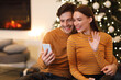 © Prostock-studio - A young couple is happily engaged in a video call, smiling and waving at the screen. They sit together by a beautifully decorated Christmas tree in a warm living room setting.