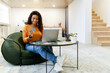 © Prostock-studio - A woman smiles as she engages in online shopping from a comfortable bean bag chair. She holds a credit card and uses her laptop in a bright, modern living space.