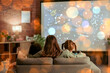 © Pixel-Shot - Cute girls with their mother watching cartoons on projector screen at home in evening, back view