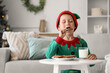 © Pixel-Shot - Cute little boy dressed as Christmas elf eating cookies with cup of milk at home