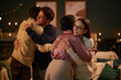 © Seventyfour - Two young adult women and two young adult men, including Black and Caucasian individuals, hugging each other in warmly lit living room, expressing happiness and connection