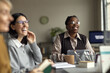 © Seventyfour - Young adult Black woman and young adult Caucasian woman sitting at table, smiling and engaging in conversation during meeting, blurred coworker in foreground, office environment visible