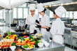 © artitwpd - Three chefs in white uniforms and hats cook in a professional stainless steel commercial kitchen, preparing dishes at the stove with fresh vegetables and ingredients arranged on the counter.