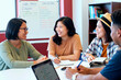 © Mediaphotos - Group of young Asian women and young Black man sitting at table discussing ideas and writing in notebooks during meeting in modern office setting with whiteboard in background