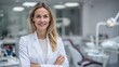 © mariodelavega - Confident female dentist standing in modern clinic, wearing white coat with arms crossed and friendly smile, blurred background with dental chair and equipment