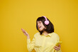 © SHOTPRIME STUDIO - A joyful young woman in a pale yellow shirt wears pink headphones and dances while listening to music on a mobile device, posed against a vivid yellow studio backdrop.
