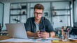 © Plaifah - Young Man in Glasses Working on Smartphone at Modern Desk with Laptop and Documents in Bright Office Space