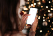 © YarikL - A woman's hands holding and touching a vertical smartphone with a blank screen, set against a blurred background of festive Christmas tree lights.