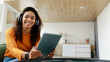 © Prostock-studio - In a well-lit room with a contemporary design, a woman with curly hair smiles as she holds a tablet. She is wearing headphones, immersed in her digital experience.