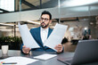 © Prostock-studio - A man in formal attire sits at a desk in a contemporary office, closely examining two papers while a laptop and a coffee cup are present nearby. The atmosphere is focused and productive.