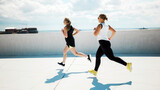 Two women jogging on a sunny rooftop with a city skyline in the background while wearing athletic attire