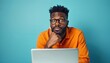 © Vadym - Confused African American man in orange shirt and glasses looks at laptop screen. Guy has worried expression, hand on chin, thinking about computer problem. Isolated on blue.