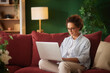 © Stockphotodirectors - A woman sits on a red couch in a bright living room, focused on her laptop. She wears glasses and a casual shirt, creating a relaxed atmosphere in the afternoon light.