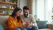 © Viktor - Happy young Indian couple smiles while looking at smartphone screen together, sitting on couch in cozy living room. Woman points at phone, sharing content with her partner during leisure time at home.