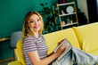 © deagreez - Young woman relaxing on a bright yellow sofa writes in a notebook at home smiling and enjoying a casual creative moment