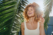 © SHOTPRIME STUDIO - smiling woman with curly hair enjoying sunny tropical outdoors, wearing white tank top framed by green palm leaves and bright sunshine