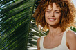 © SHOTPRIME STUDIO - smiling woman with curly hair standing near tropical palm leaves, wearing white tank top, natural sunlight illuminating her face outdoors, warm summer vibe