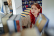 © Seventyfour - Caucasian teenage girl with red hair reaching for book on library shelf, focused expression on face, visible piercings and backpack, surrounded by blurred books in foreground