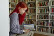 © Seventyfour - Teenage Caucasian girl with long red hair sitting in library reading book, holding open hardcover while focusing on pages, surrounded by shelves filled with books in background