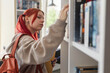 © Seventyfour - Teenage Caucasian girl with red hair browsing bookshelf, holding notebook and pen, wearing backpack, standing in library or bookstore, searching for book, focused expression