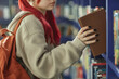 © Seventyfour - Teenage Caucasian girl with red hair selecting book from library shelf, wearing backpack, showing black painted fingernails, standing among rows of books, face partially visible