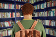 © Seventyfour - Back view of Black teenager wearing backpack and headphones standing in front of bookshelves in library, appearing to listen to audio or music while looking at book selection