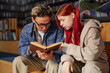 © Seventyfour - Teenage Caucasian girl with red hair and Asian boy sitting together reading book, both focused on pages, sharing learning experience in library setting
