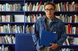 © Seventyfour - Portrait of Asian young adult man with short hair and glasses standing in front of bookshelf holding blue folder, looking directly at camera in library setting