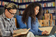 © Seventyfour - Young adult Asian man reading book while young adult Black woman working on laptop in library setting, both focused on tasks, bookshelves visible in background
